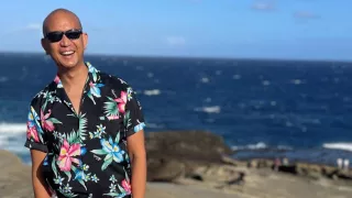 A man in a Hawaiian shirt standing on a rocky beach with the ocean in the background
